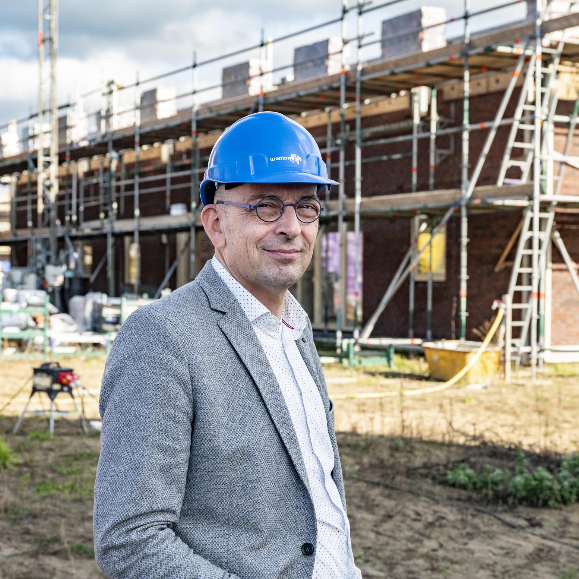 Gerrolt Ooijman (Wonion) in pak met blauwe helm voor een bouwplaats met huizen in aanbouw